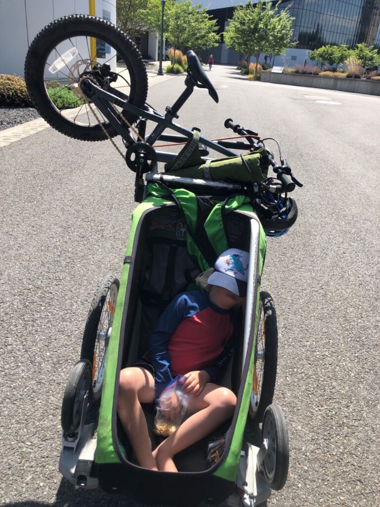 Child napping in a green bicycle trailer with child's bike attached to the back of trailer.