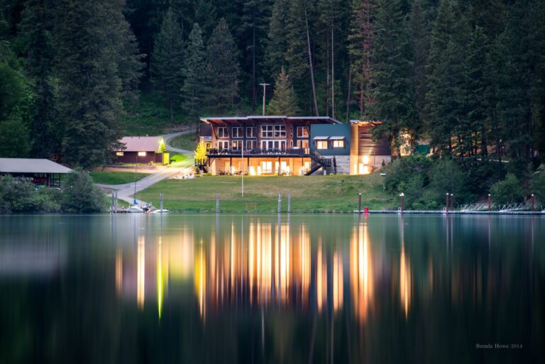 Main lodge at Camp Four Echoes at dusk with all the lights on and reflection of lodge and lights on the calm, flat surface of Lake Coeur d'Alene.