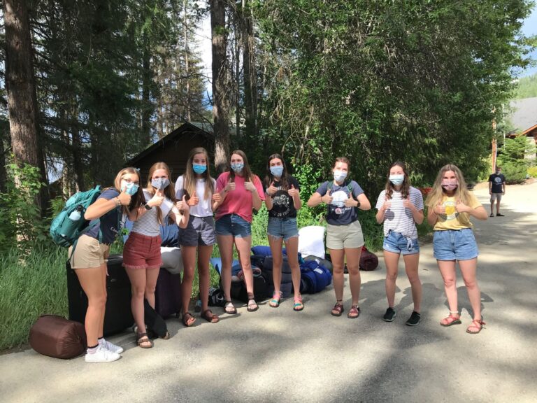Happy teenage girls at Camp Spalding during summer 2020, gathering in a line, looking at camera, smiling behind their COVID face masks, and giving the camera thumbs-up signs. Trees in the background.