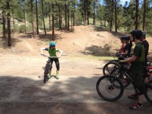 Dirt area of a park with a mountain biking instructor demonstrating a skill with a bike to a group of students.