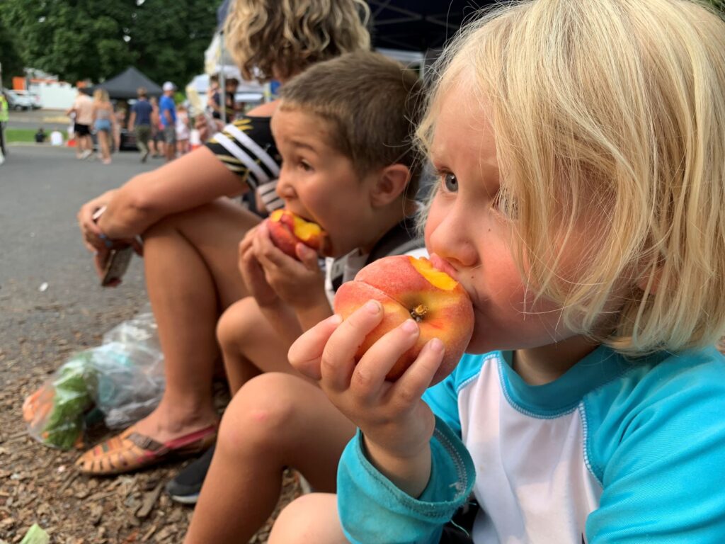 Two children sitting down and eating fresh peaches at a local farmers market.
