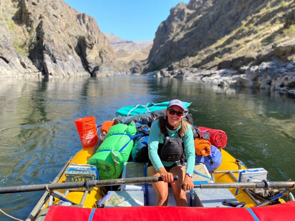 Female river raft guide, wearing a life jacket, sunglasses, and hat, sitting in a raft surrounded by colorful gear bags, with the river and rocky riverbank in the background.