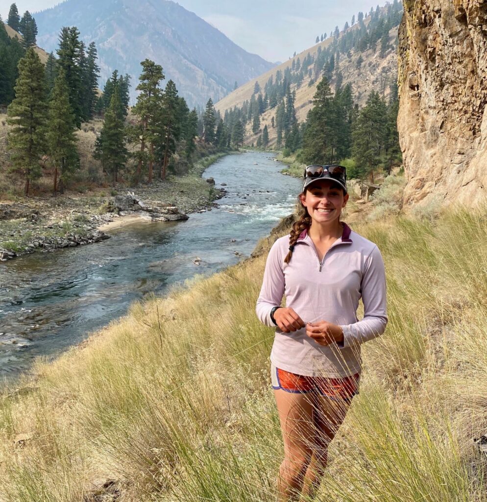 Woman smiling at the camera, standing in a grassy riverbank, with view of river, trees, and hillsides in the background.