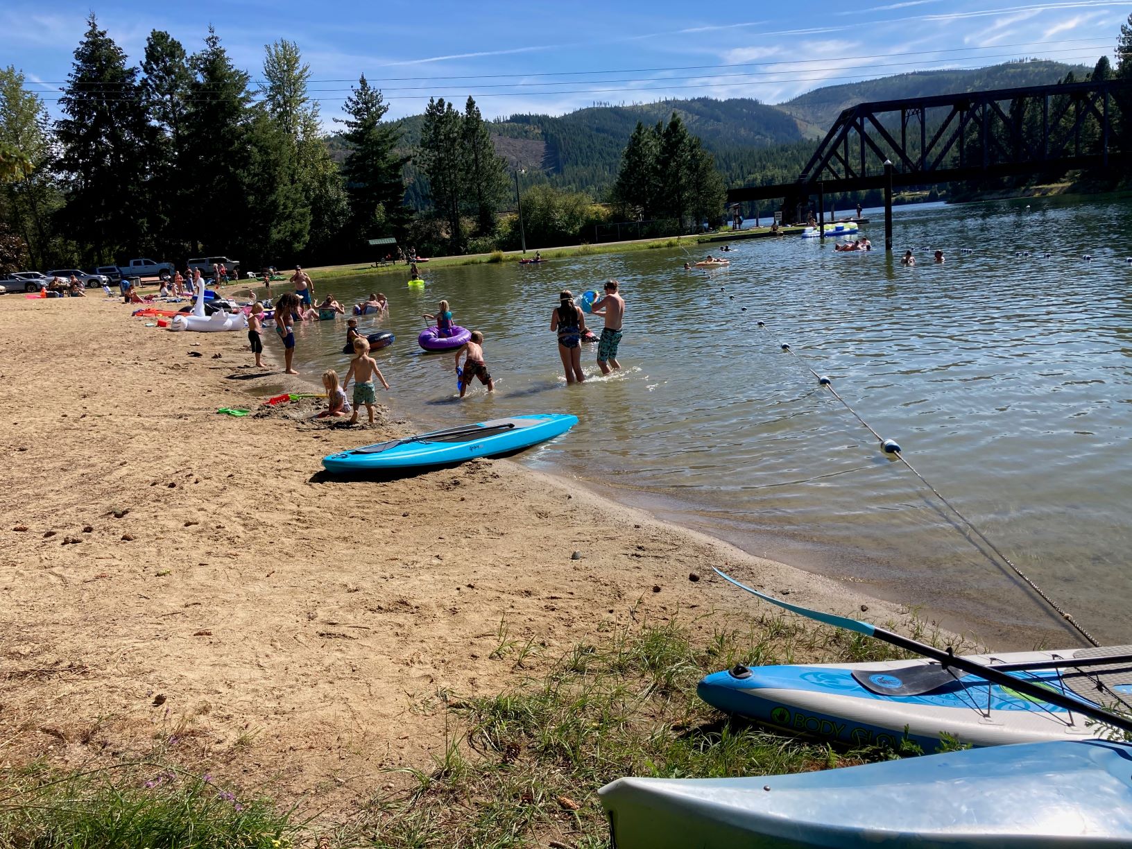 People standing on the sandy beach and in the water at Priest River Recreation Area, with kayaks and other paddling gear on the shoreline.