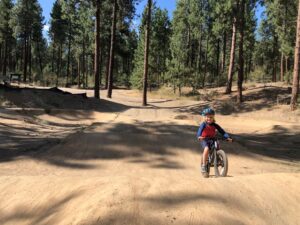 Young child riding his bike on a dirt pump track at Camp Sekani, with Ponderosa pine trees in the background.