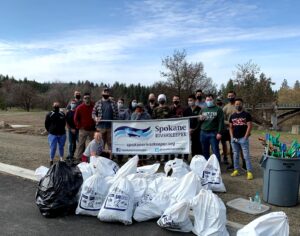 Group of volunteers for the Spokane River Clean-Up, holding a sign that says Spokane Riverkeeper, and nearly 20 huge plastic bags of trash, depicting how much debris and litter was cleaned up.