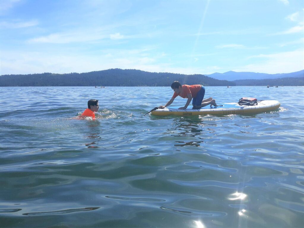 Two boys swimming in Lake Coeur d'Alene, one boy kneeling on a paddleboard. Sun shining on the water, and mountainous hills in the far background.