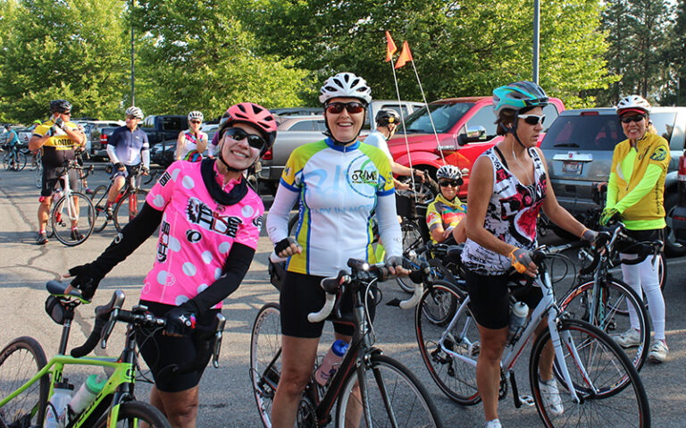 Two women on bikes, wearing clothing gear and helmets, smiling at the camera, surrounded by other cyclists during Spokane Valley's Cycle Celebration.