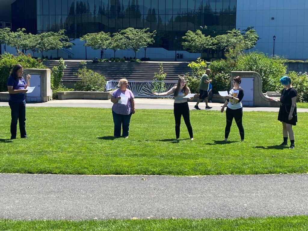 Actors standing on the grass rehearsing for Shakespeare in the park.