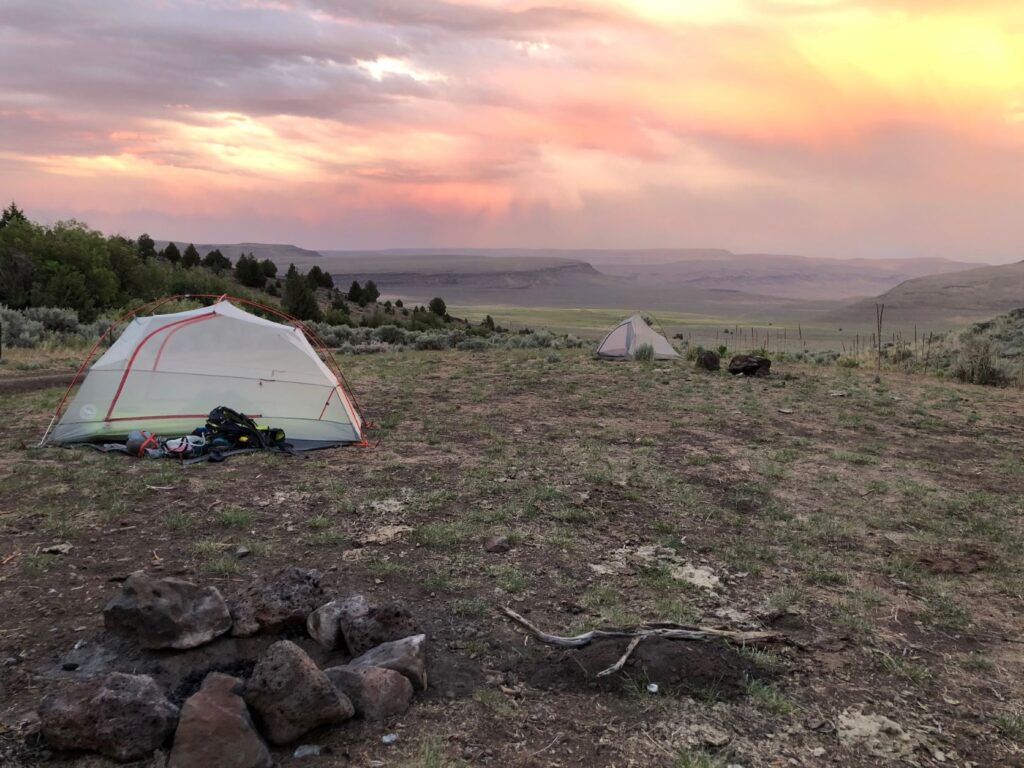 Tents in a canyon on the east of Steens Mountain, with a yellow, pink, and purple sunset in the background.