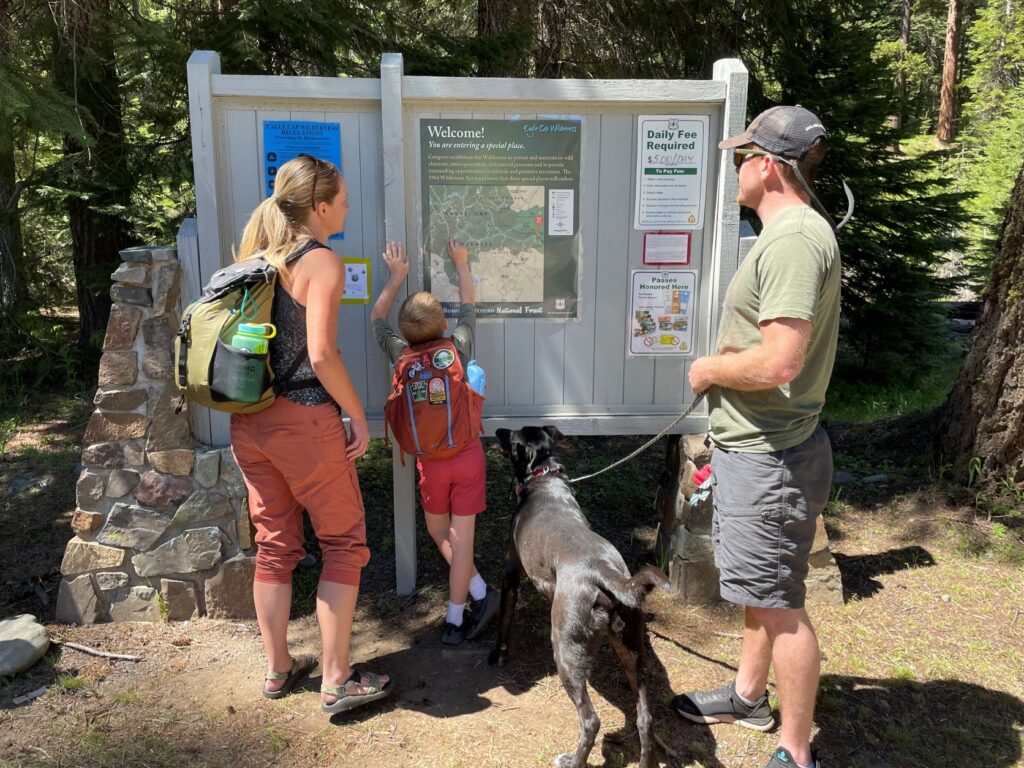 Child reading a trail map at a trailhead kiosk with two adults looking on.