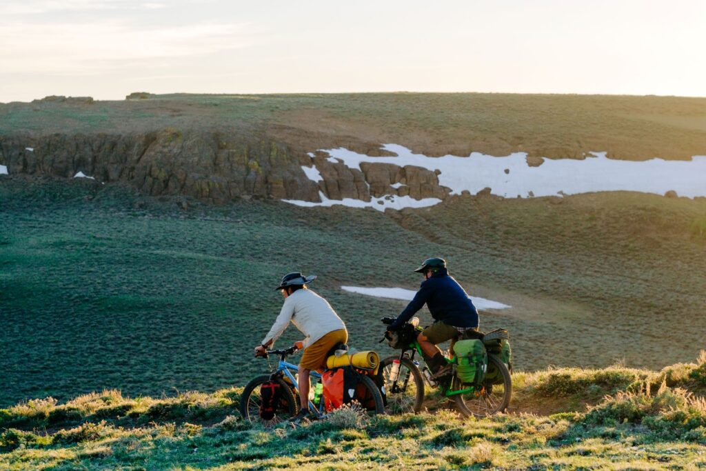 Two bikers along a dirt trail in te Trout Creek Mountains, with snow remnants on the shady rock cliffs in the distance.