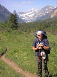 Writer backpacking in Glacier National Park, on a singletrack trail through a meadow with a mountain peak in the distance.