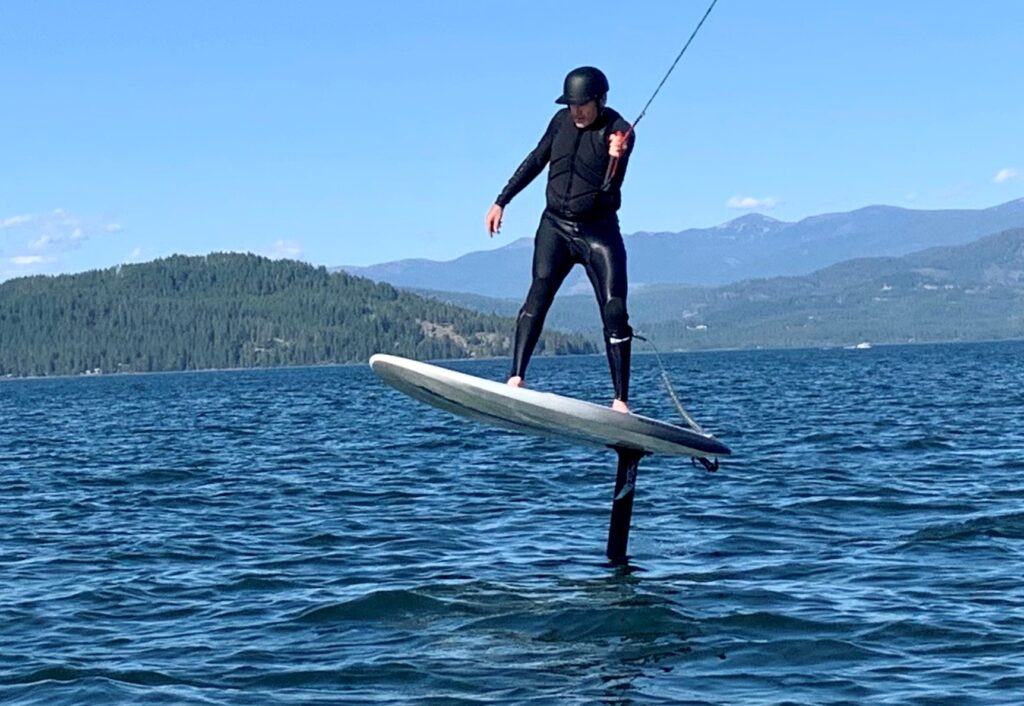 Man standing on wind foiling board, getting towed behind power boat, on Lake Pend Oreille.