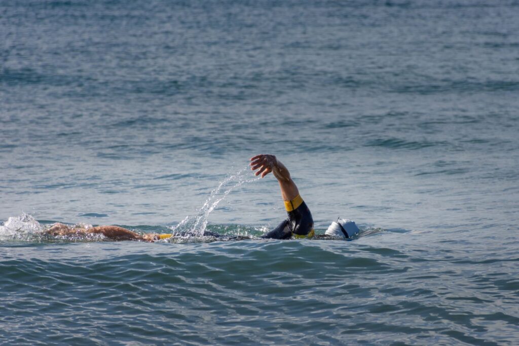 Swimming doing freestyle strokes in open water, wearing a wetsuit, swim cap, and goggles.