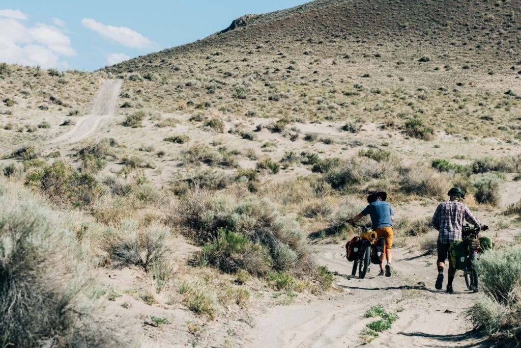 Two bikers pushing their bikes, loaded with bikepacking gear, through the sadn drifts of Big Sand Gap in Oregon.