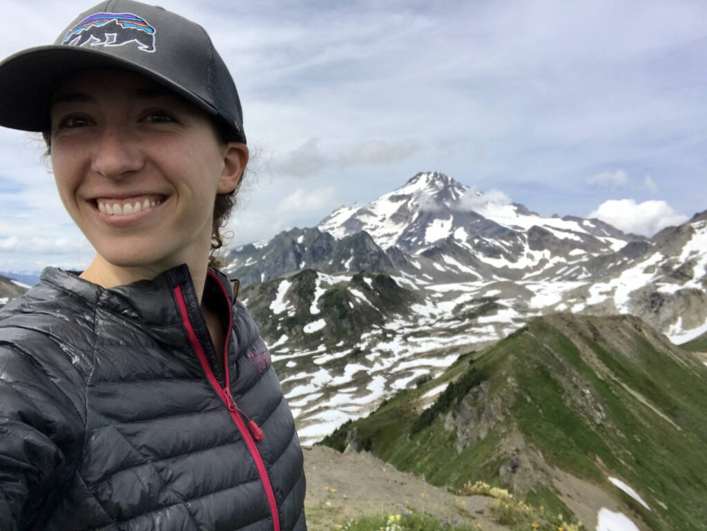 Jade Tabony smiling atop a hiking ridge with a snowy mountain peak in the distance.