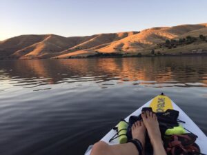 Paddler's view of the glowing Snake River, during sunset, with the sloping hills and paddler's feet resting on her paddleboard.