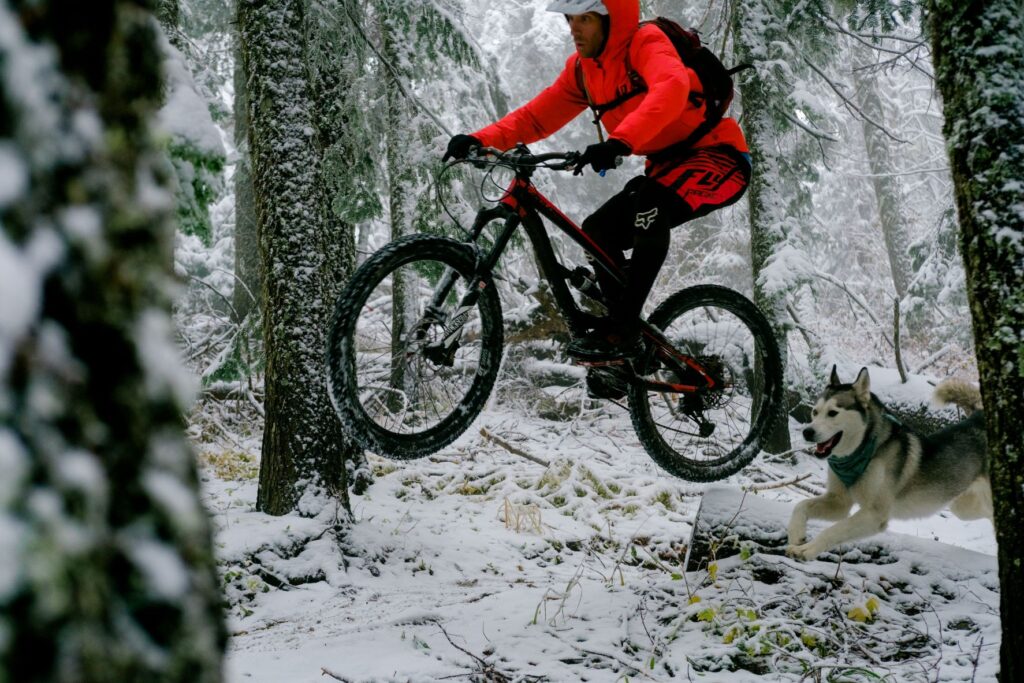 Mountain biker getting air riding off a jump along a snowy trail with their husky dog gleefully running along.