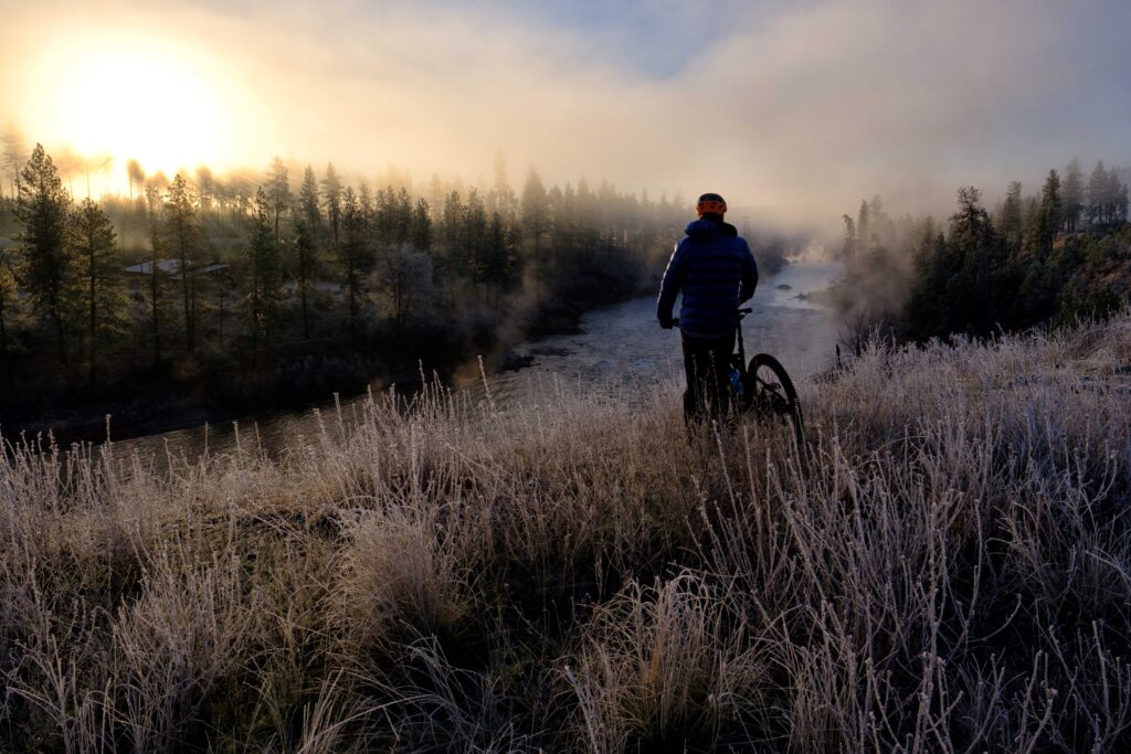 Cold, clear mornings on the Spokane River often bring sun, steam, and frost, as well as the opportunity to ride in a puffy. Biker is stopped along a trail gazing in the distance at the Spokane River below and forested hillside across the river.