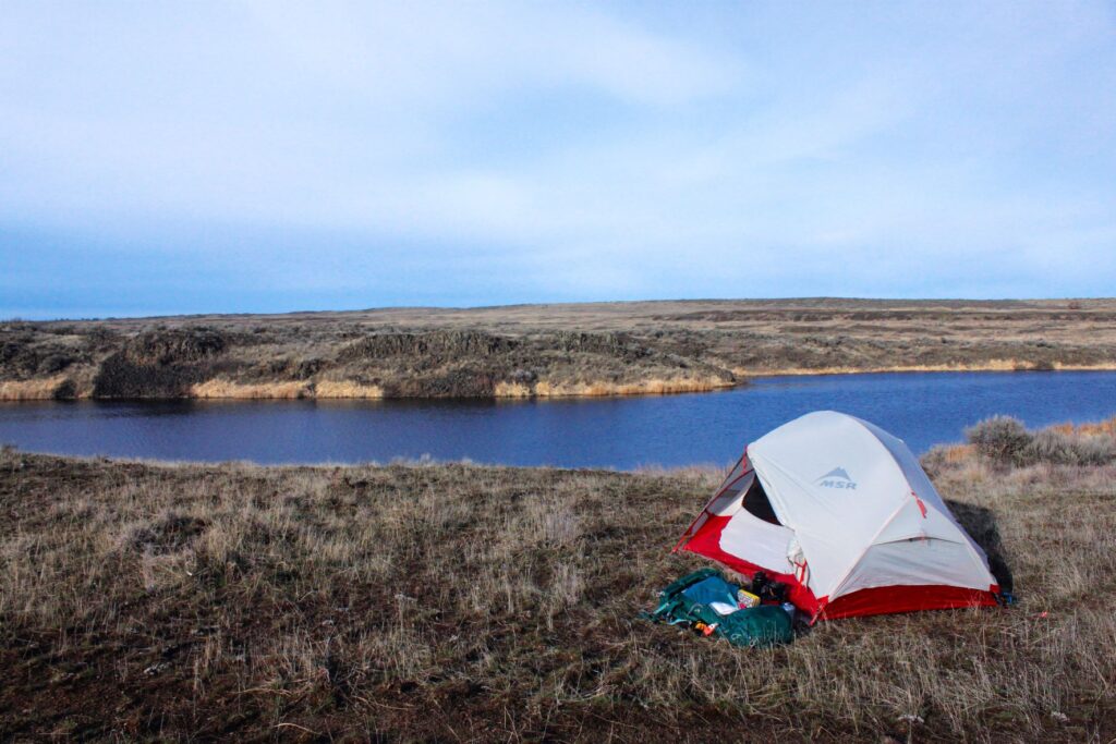 Small white and red tent in an open patch on wild bunch grass near Z Lake, with flat topography in the distance and party cloudy blue sky.