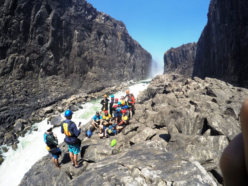 Group of boaters in their paddling gear and helmets sitting on the rocks along the Zambezi River below Victoria Falls. /