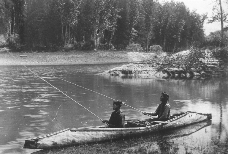 Sepia-tone photo of traditional Kalispel Tribe canoe and two people sitting inside with fishing rods in the water.