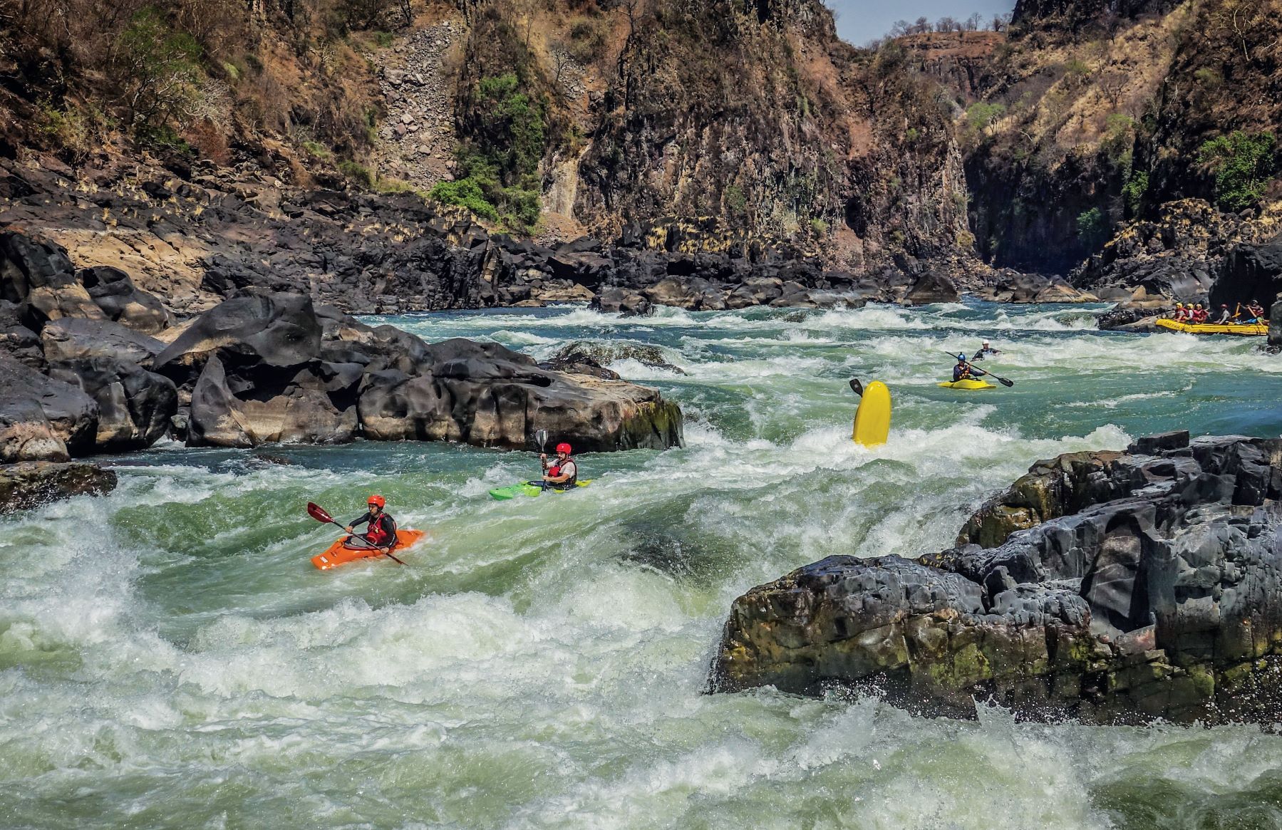 Group of kayakers paddling the big whitewater rapids on the Zambezi River, with one kayak tip high up, and a group of people in a raft upriver.