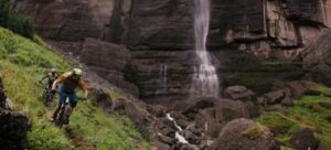 Two people riding on mountain bikes on a singletrack trail by a waterfall.