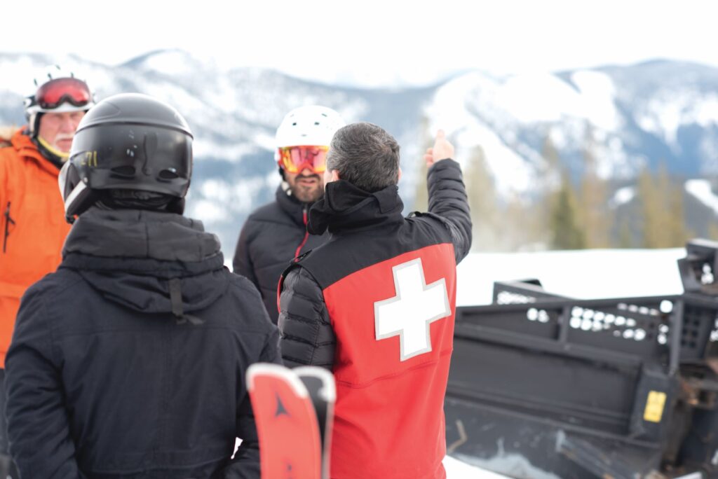 Skiers gathered together to listen to a safety briefing led by a ski patroller, wearing a red ski jacket with a big white cross on the back.