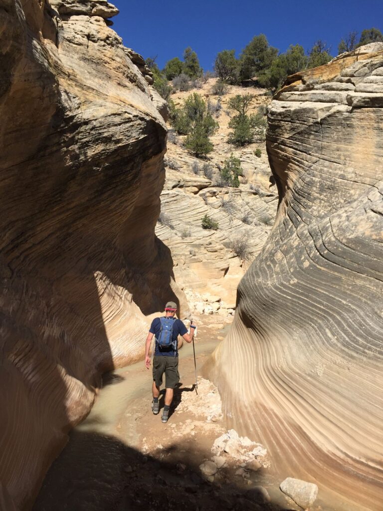 Man hiking through a sandstone slot canyon in southern Utah, with high canyon walls and narrow opening.
