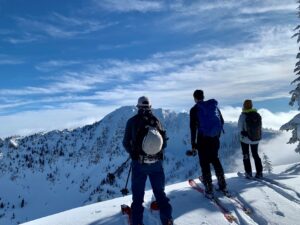 Three backcountry skiers, backs to the camera, looking over the edge, preparing to ski down, with a snowy ridge line farther beyond.