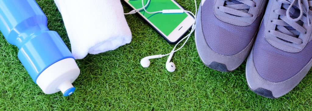 Scene looking down at grass with a water bottle, rolled-up white towel, smart phone with earbuds, and pair of running shoes.