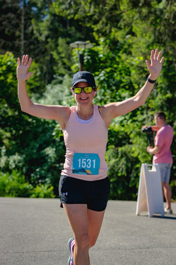 Woman running in the Coeur d'Alene Marathon with arms up in celebration, smiling at the camera.