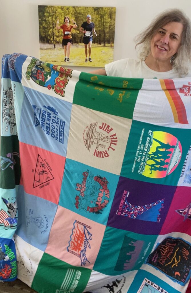 Author holding her quilt made from the graphic front side of old race t-shirts--standing in her living room with a canvas photo print of her running with her wife.