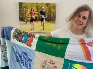 Author holding her quilt made from the graphic front side of old race t-shirts--standing in her living room with a canvas photo print of her running with her wife.