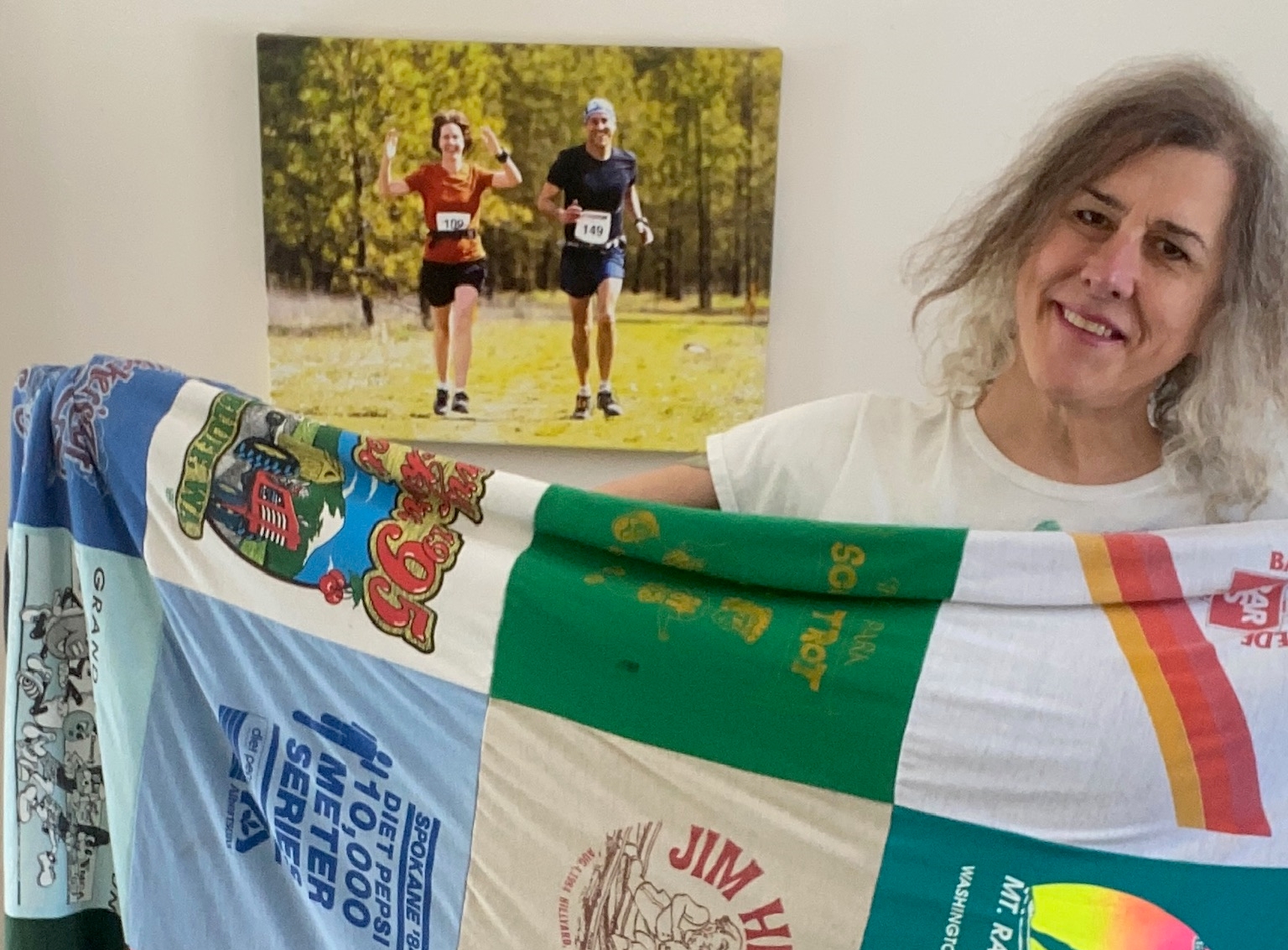Author holding her quilt made from the graphic front side of old race t-shirts--standing in her living room with a canvas photo print of her running with her wife.
