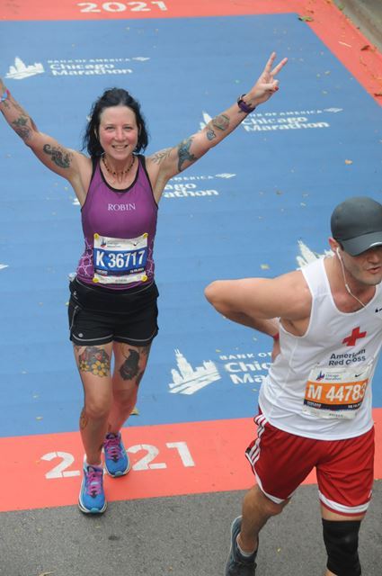 Robin Manees running across a marathon finish line with both arms raised high, fingers with 2-finger peace signs, in triumph