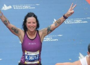 Robin Manees running across a marathon finish line with both arms raised high, fingers with 2-finger peace signs, in triumph