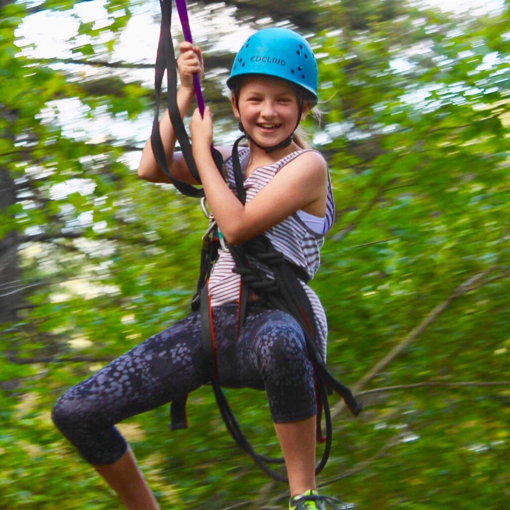 Child smiling while zooming down a zipline, wearing a blue helmet and safety harness, with forested background.