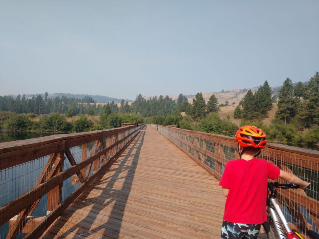 Young boy wearing a red shirt and orange bike helmet is standing next to his bike on a long brown wooden bridge that's part of the Ferry County Rail Trail.