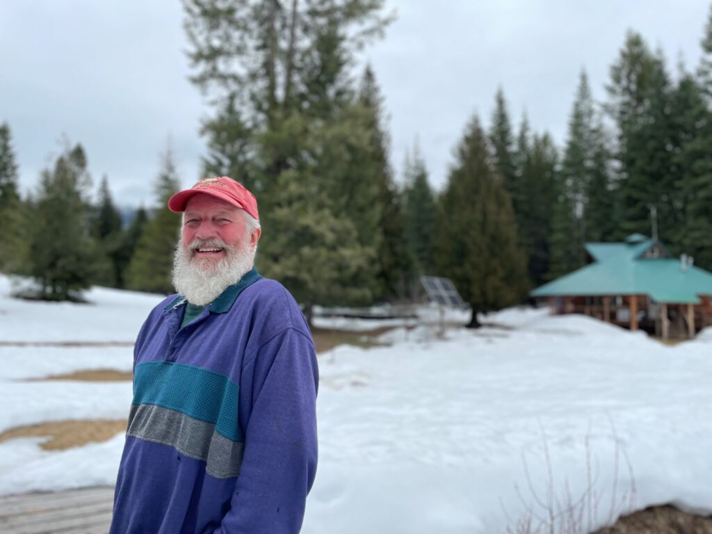Man named Fish is standing angled to the side looking and smiling at camera, and in the background is snow-covered ground and pine trees, with a view of his cabin to the right.