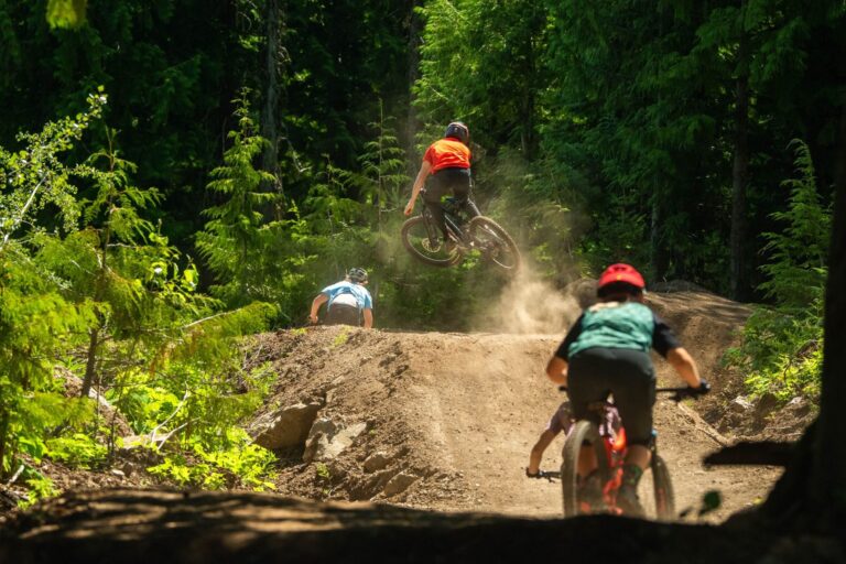 Four mountain bikers, view from behind, as they ride up and over a dirt jump hill, with one rider catching air at the top.