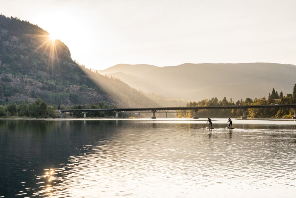 View of Arrow Lakes region and two standup-paddlers going across the water with a long bridge in the background and hills, with the sun setting behind them.