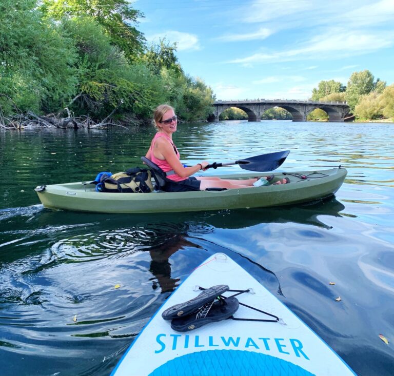 Woman smiling while kayaking on the Spokane River, looking back at her friend that's on a Stillwater brand stand-up paddleboard -- bridge over the river in the distance.