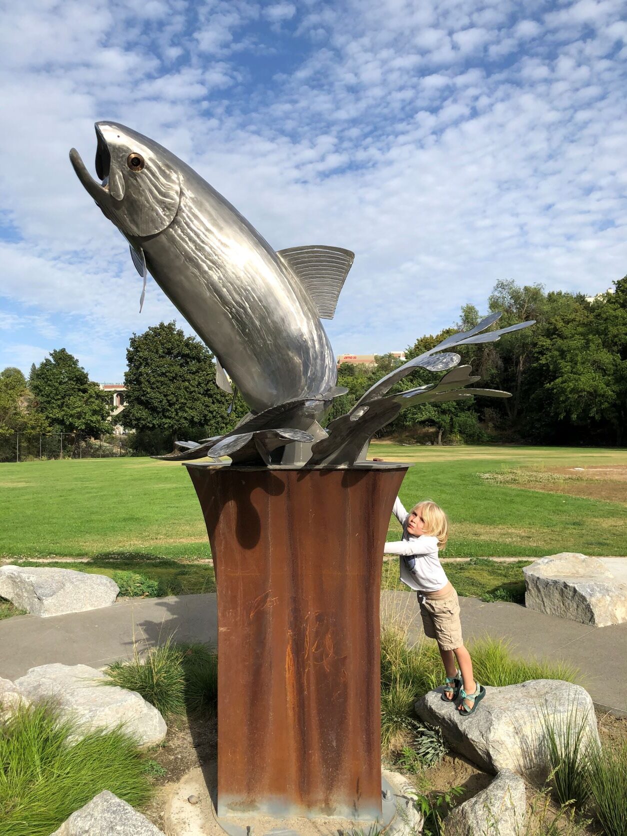 Young boy leaning against the wooden base displaying metal sculpture of redband trout at Redband Park in Spokane.