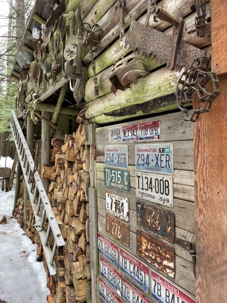 Outer wall of log cabin owned by a man named Fish is decorated with vehicle license plates from different states and rusted metal tools and other artifacts that Fish has found around his property. A high stack of firewood is next to the cabin.