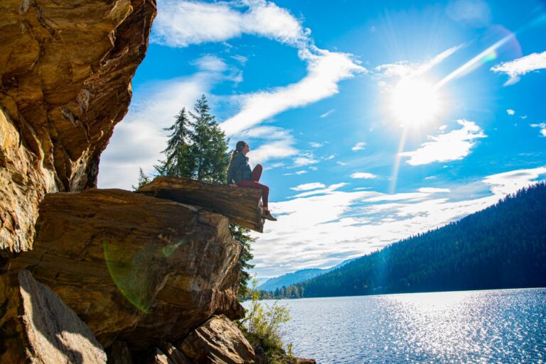 Female hiker sitting on a log resting overlooking Slocan Lake with trees down below and blue sky above.