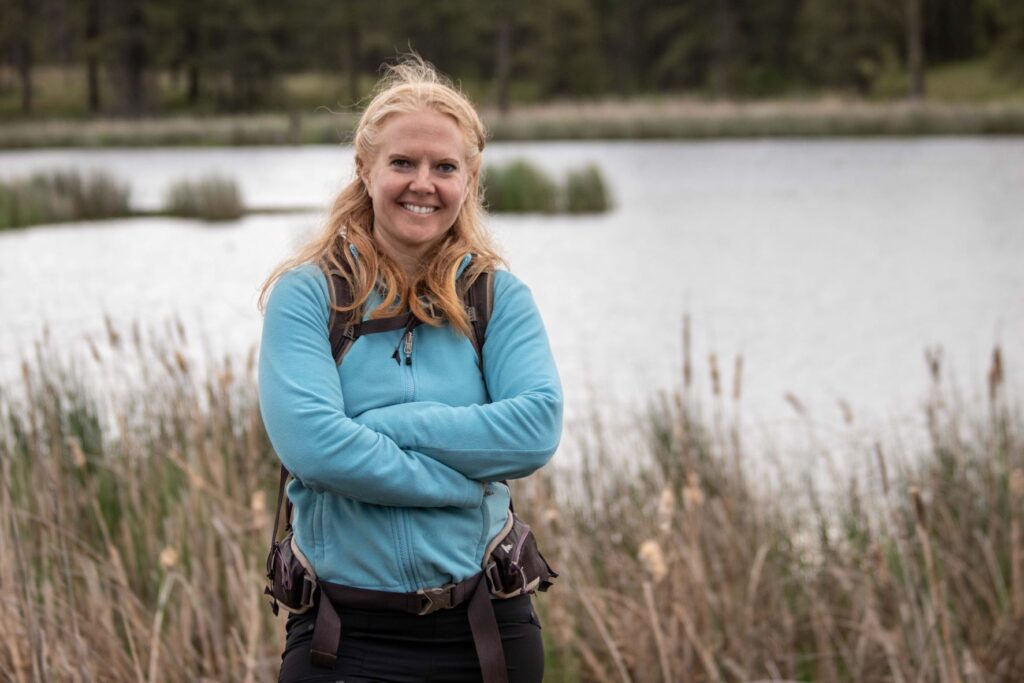 Gina Claeys standing wearing a blue long-sleeved shirt and backpack with view of lake and grassland in the background.
