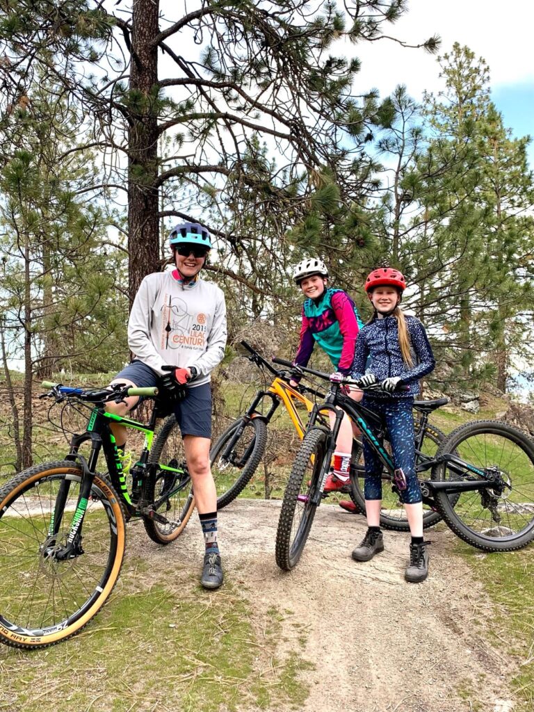 Adult and two youth sitting on their mountain bikes and smiling at the camera along a forested dirt trail.